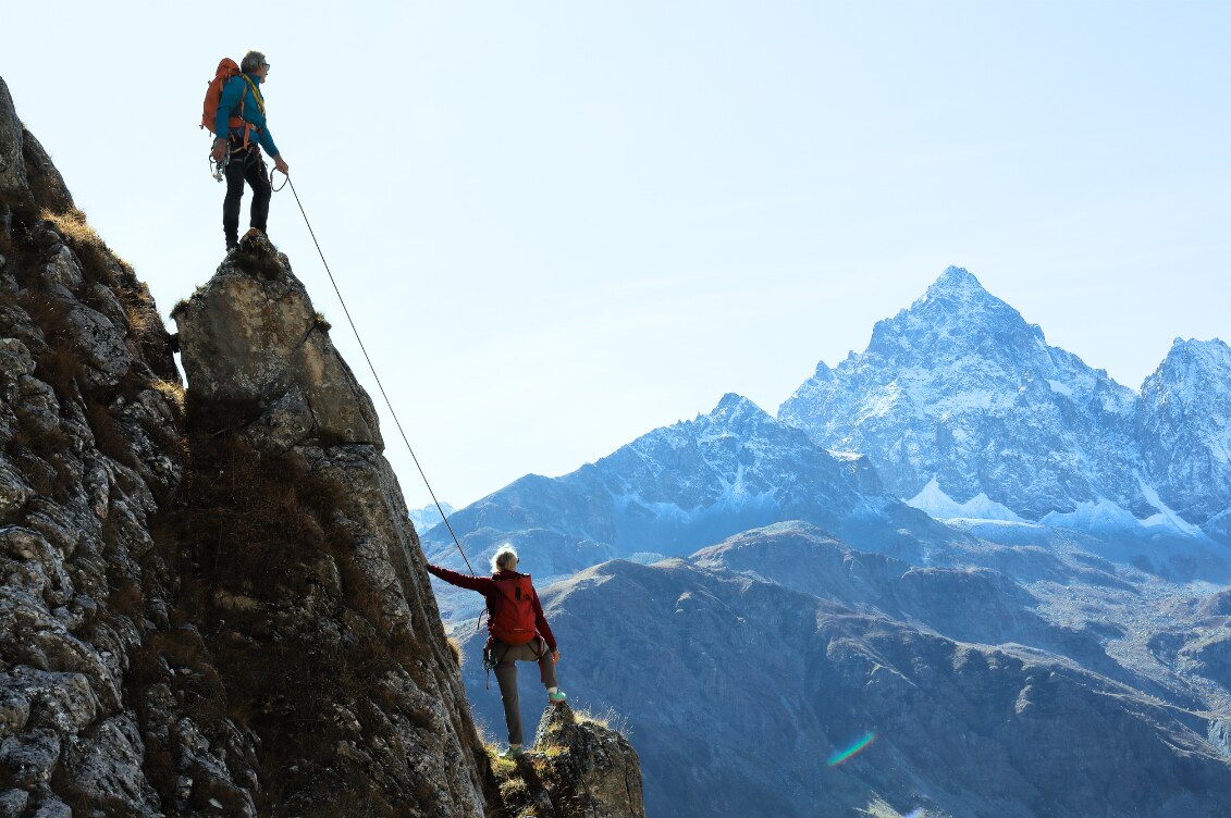 Two climbers roped together on a mountain ridge, one reaching back to guide the other up