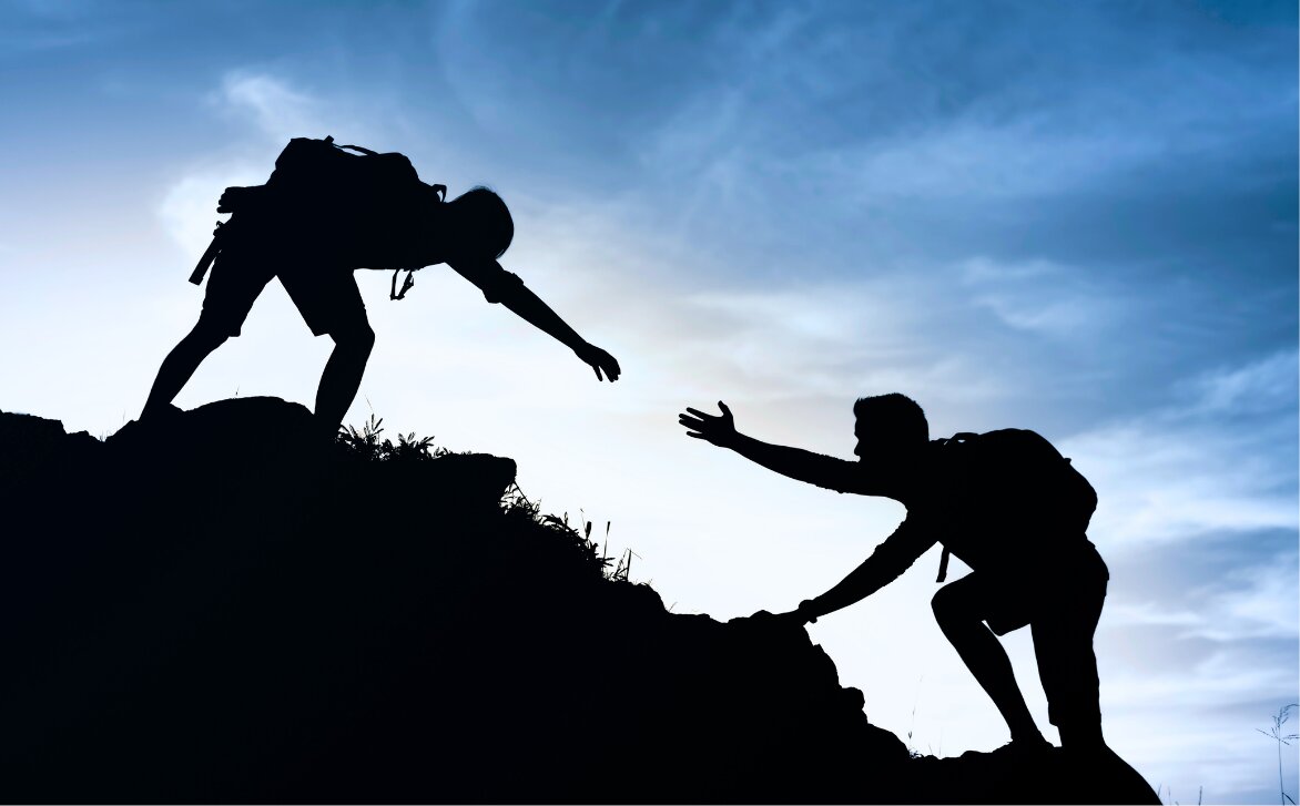 One hiker reaching down to help another climb up a ridge